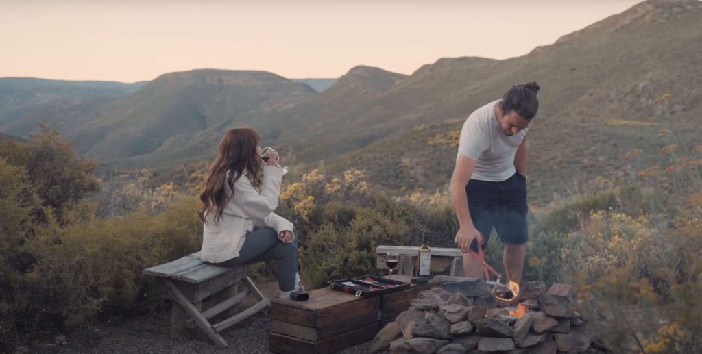 A couple preparing a meal and having a glass of wine at their campsite on high ground. 