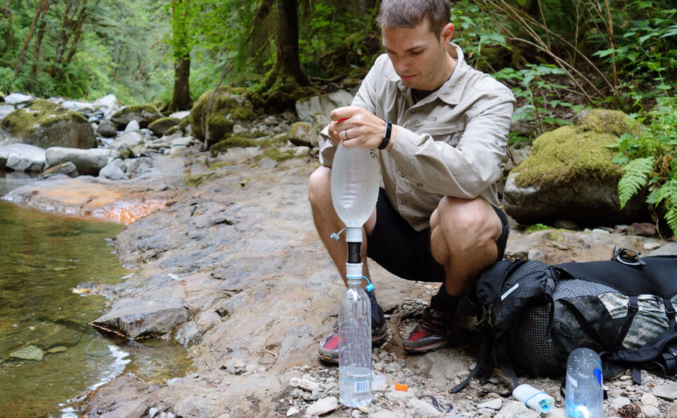 A man backpacking by the river, squatting down to fill his HydroBlu Versa Flow water filter with river water.