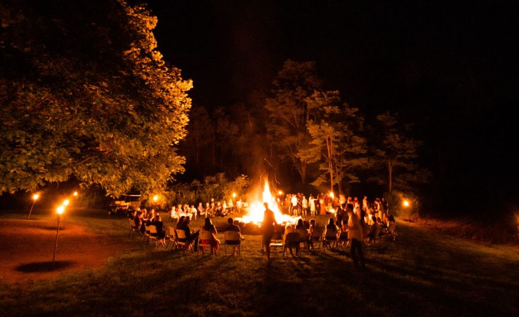 A large group of people sitting around a blazing campfire at night waiting for a story.