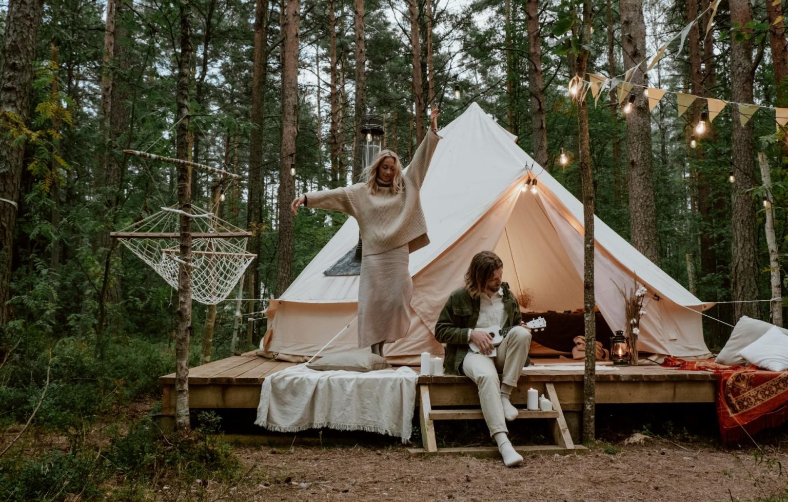 Couple glamping. Man is playing a ukulele. A hammock is strung up next to their glamping tent.