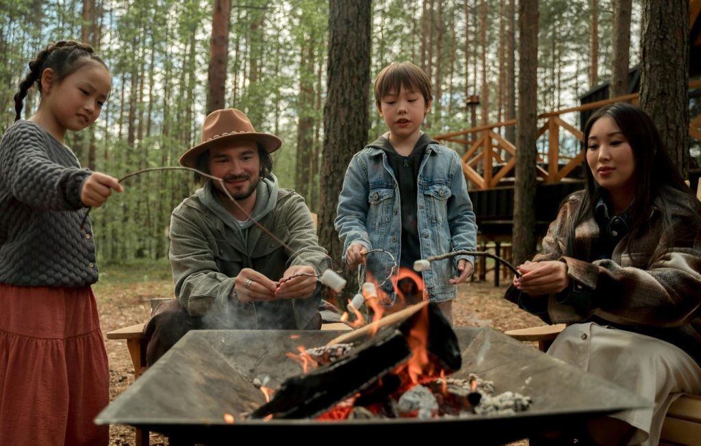 Family camping builds lasting memories. Husband, wife, son and daughter are roasting marshmallows over a fire pit in front of their glamping cabin.