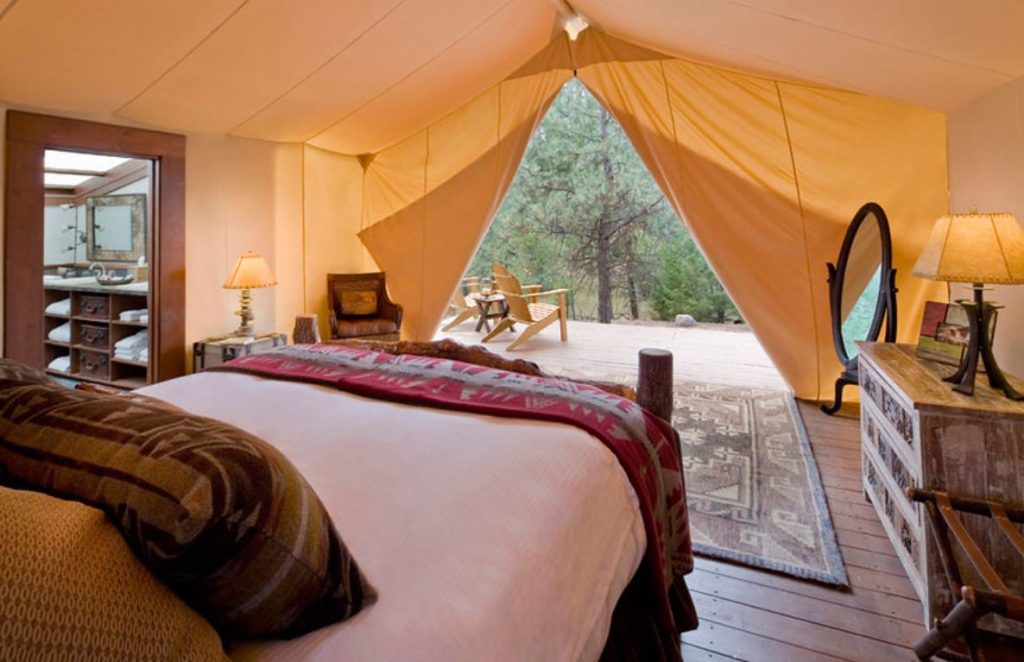 Inside of a glamping tent showing comfortable bedding and very nice amenities throughout. Tent flap is open to a forest view.