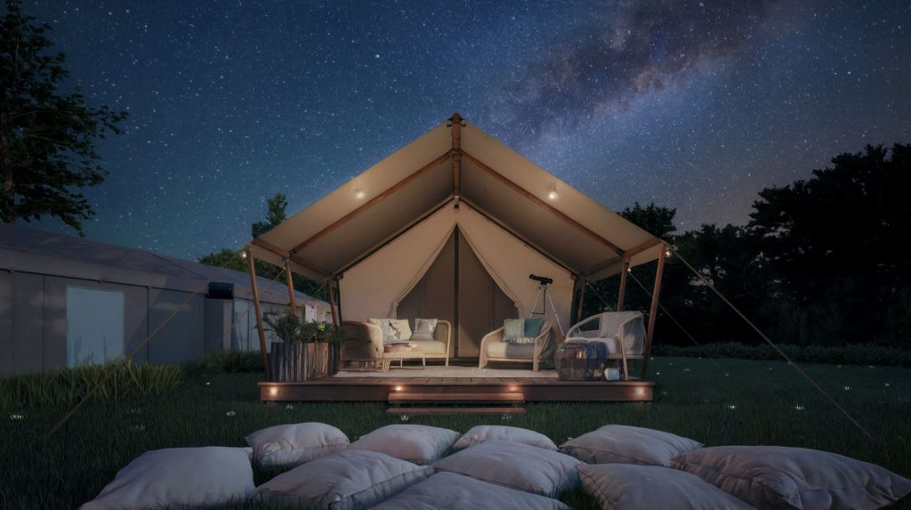 Luxury glamping tent under the stars. The tent has a porch with a telescope and other outdoor furniture. The background reveals a clear night sky filled with stars and a few trees. The ground is covered with soft, white cushions.