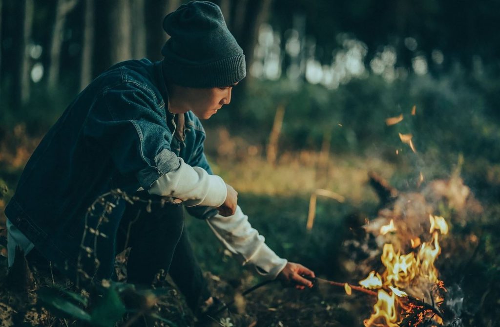 Man in a beanie by a campfire at night putting a stick in with hand way too close to the fire.