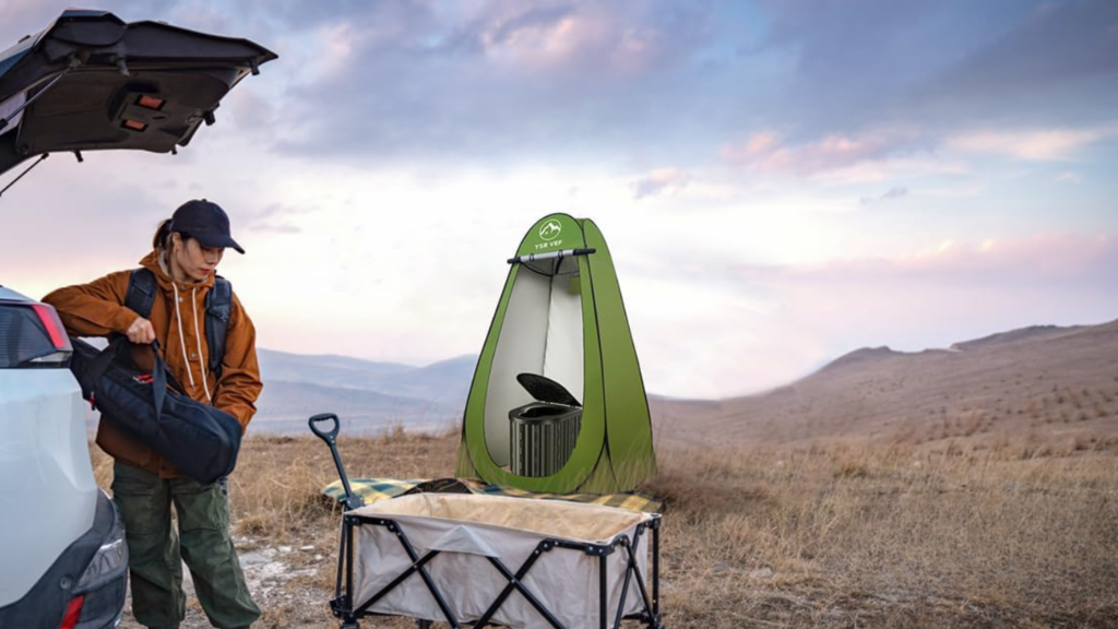 A portable camping toilet with privacy tent behind a woman unloading other camping gear in a wide open mountain field.