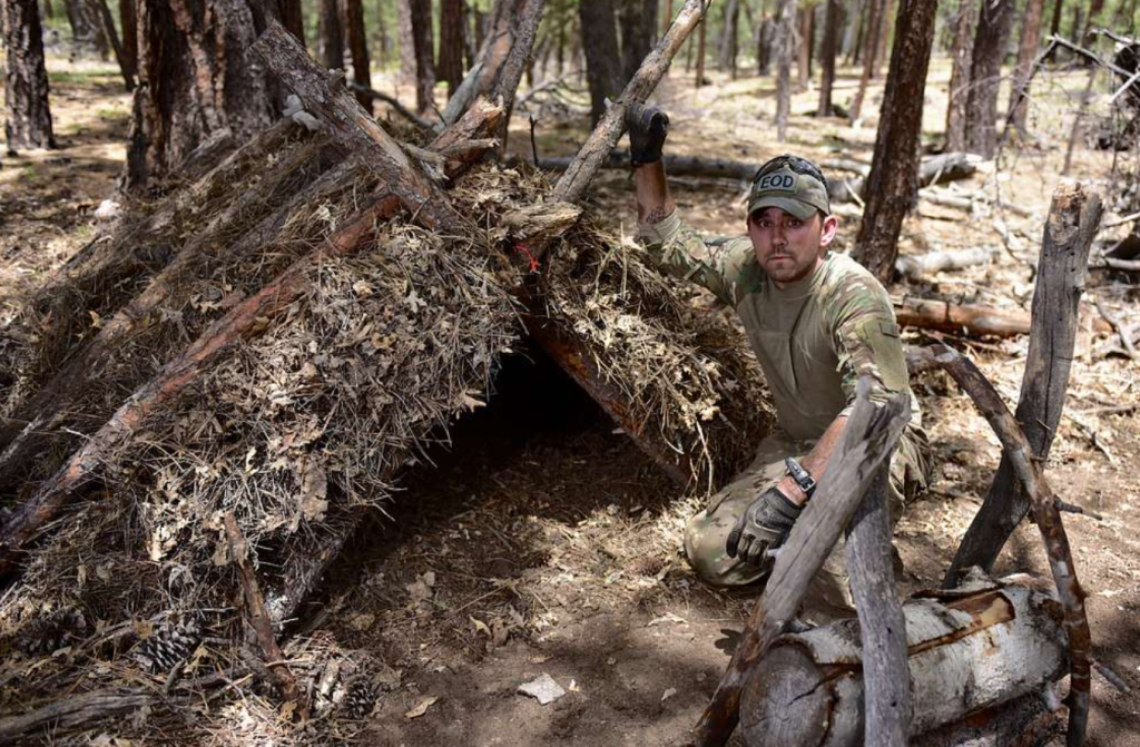 A man poses with his completed A-frame survival shelter, built for durability and warmth.