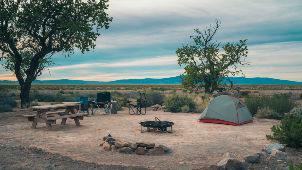 A photo of a typical campsite found at Bureau of Land Management sites. There is a tent pitched on a flat piece of land near a single fire pit and a picnic table. A few camping chairs are placed near the table. There's a tree nearby. The background is a vast, open landscape with mountains in the distance. The sky is cloudy.