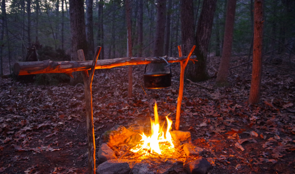 A campfire is shown toward evening, set up away from an A-frame survival shelter for campsite safety.