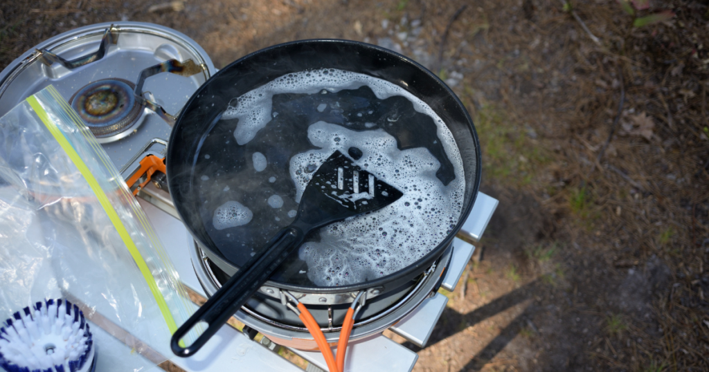 A skillet with utensils being cleaned with biodegradable soap at a campsite.