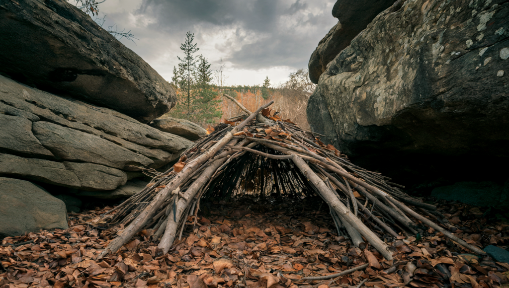 A photo of a debris-hut survival shelter in the wilderness. The shelter is built using fallen branches and leaves. It is nestled between two rocks, with the rocky terrain continuing behind the shelter. The ground is covered with dead leaves. The sky is overcast, with a few trees visible in the background.