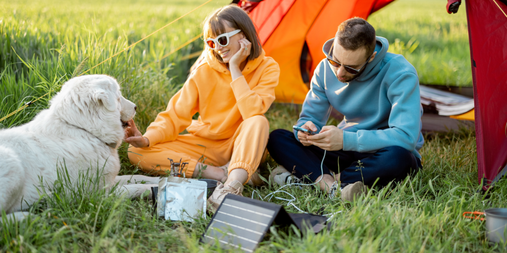 A couple is sitting on the ground with their pet Newfoundland, charging a phone in front of their tent with a flexible solar panel charger.