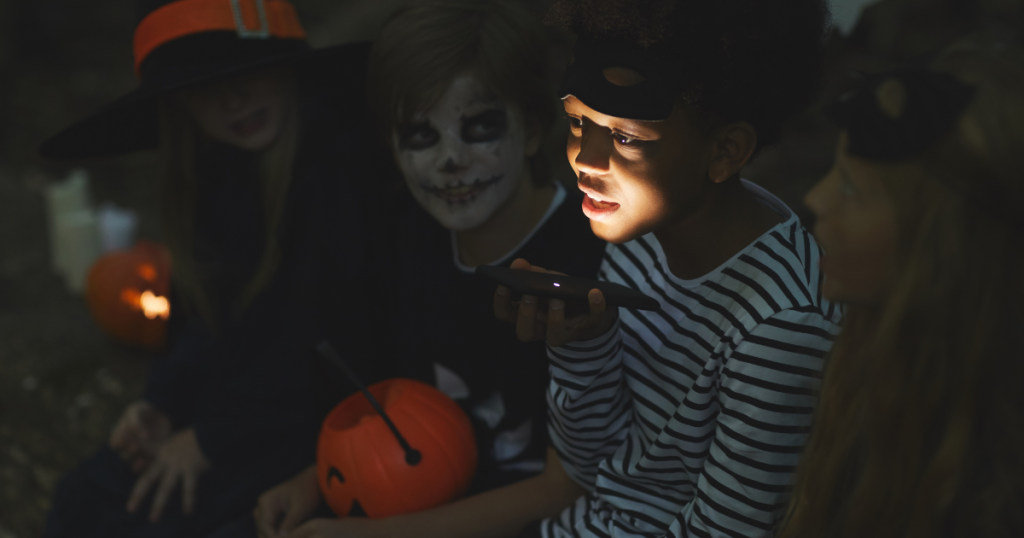 Two girls and two boys sitting around a campfire wearing Halloween costumes for spooky season.