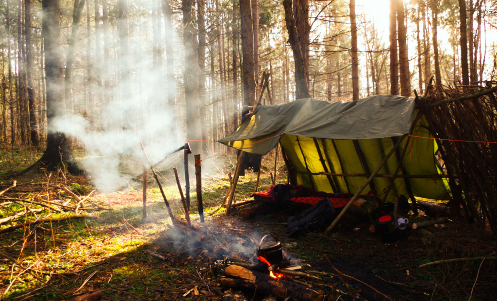 A simple survival shelter for camping made with a tarp strung between branches gathered in the wilderness. A small campfire is seen in front on a sunny, cold afternoon.