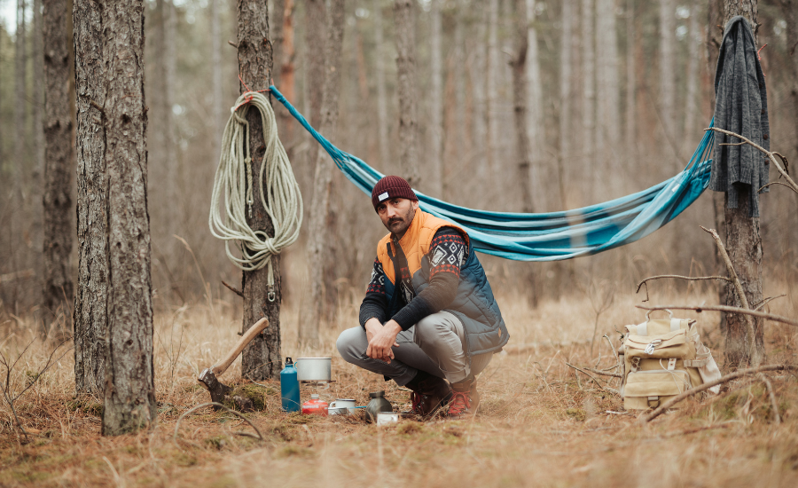 A man is seen selecting a good location between large trees to set up a survival shelter. He has a hammock strung up temporarily, a rope, a hatchet and an assortment of basic survival gear.