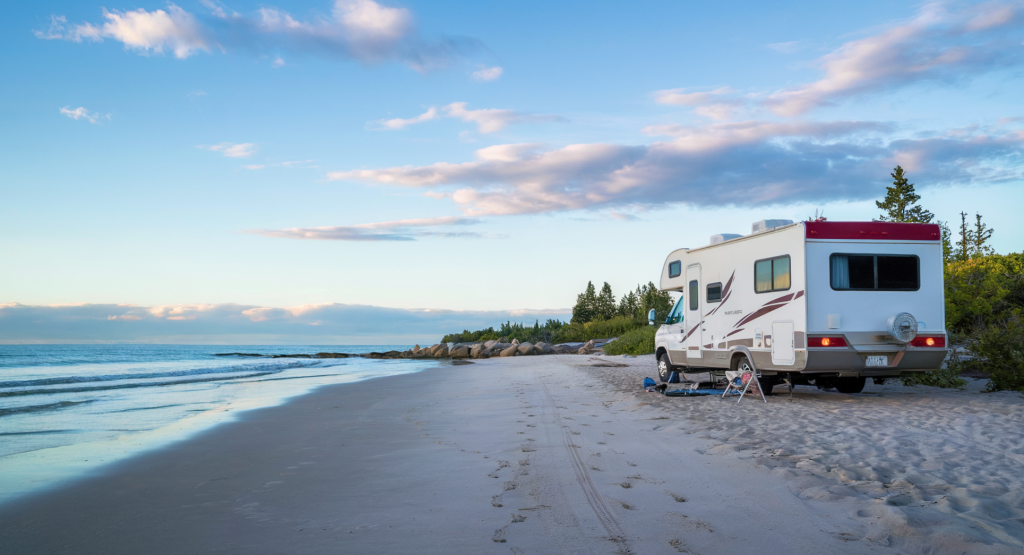 A photo of a mid-size RV parked along a remote beach. The RV is white with a red roof and has a few personal belongings outside. The beach is clean and has a few footprints leading to the water.  