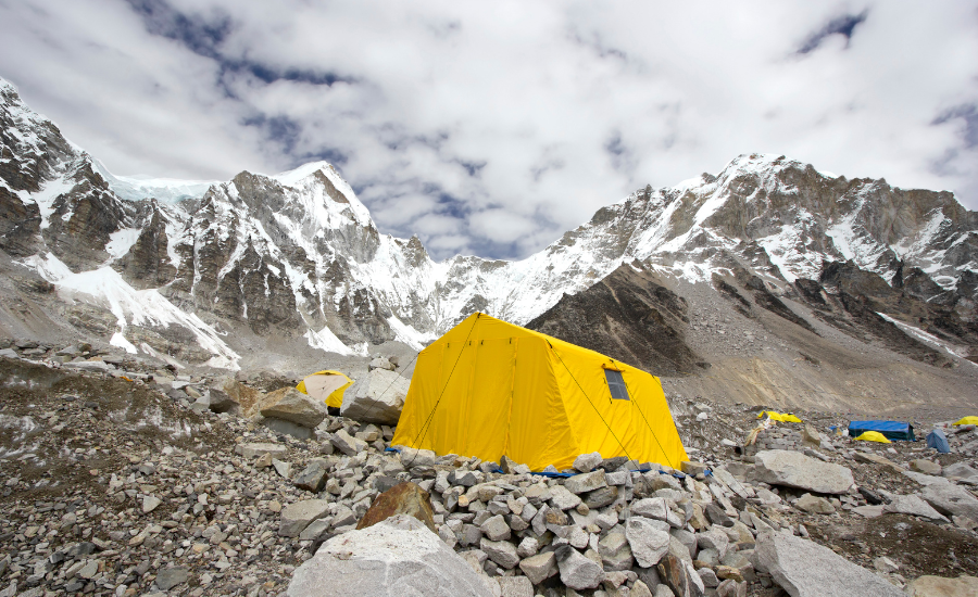 A large tent set up in a high-altitude snowy mountain. There is a natural windbreak from the mountains, and a reflective rock wall that has been built next to the tent.