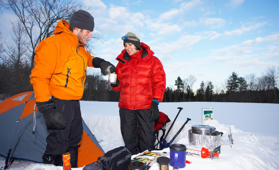 A couple sharing a hot drink from a thermos. Other food and drink gear, like a backpacking stove can be seen near their tent.