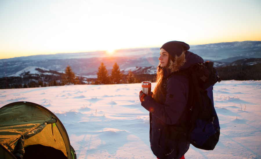 Woman dressed in layers for cold weather camping, she's wearing a backpack and holding a thermos. There is a camping tent to the left side.