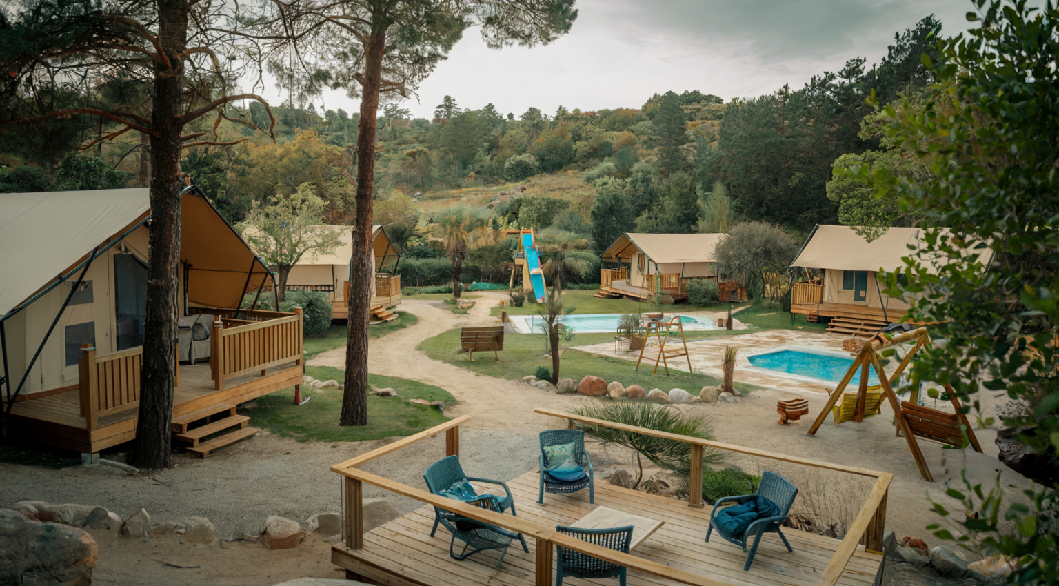 A photo of a glamping site with multiple tents, some with wooden decks, surrounded by lush greenery. There are tall trees and a dirt path leading to the tents. In the foreground, there's a wooden deck with a few chairs and a table. The site has multiple facilities, including a playground with a slide and swings, a pool, and a few benches. The atmosphere is serene, with a few trees and rocks scattered around the site.