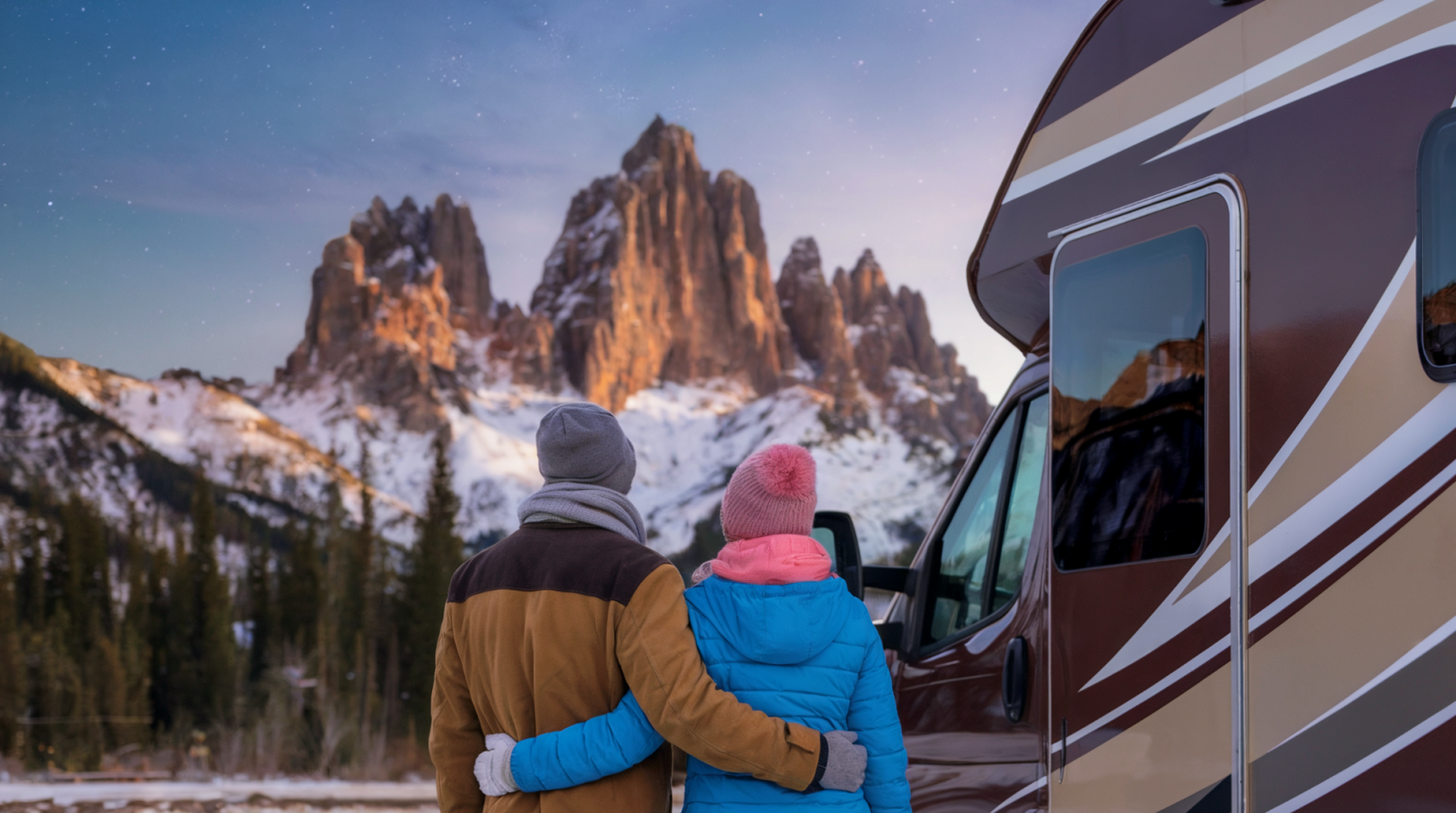 A couple wearing winter gear, standing next to their RV looking at a magical snowy mountain scene. The man is wearing a brown coat, a gray scarf, and a gray beanie. The woman is wearing a blue coat, a pink scarf, and a pink beanie. The RV has a brown and beige exterior. The snowy mountain scene in the background has tall, jagged peaks covered in snow, with a few trees near the base of the mountains.