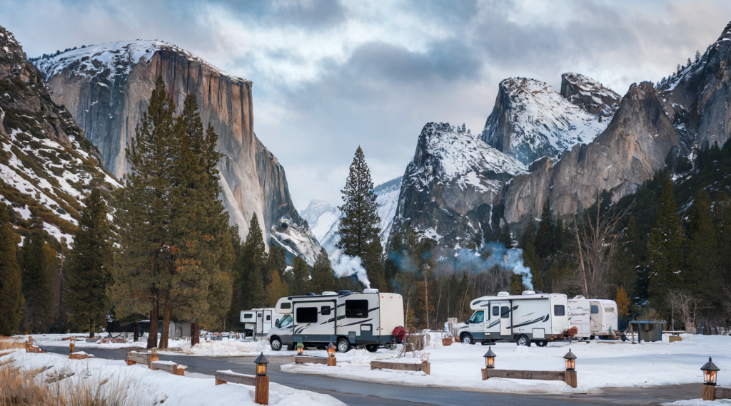 Small RV park in beautiful snowy mountains . The RV park is surrounded by tall trees and snow-covered mountains. There are several RVs parked in the park, and some have smoke coming out of their chimneys. 