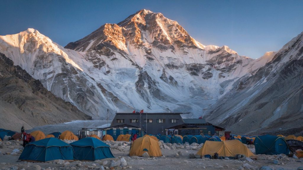 A photo of the Mount Everest Base Camp with a few tents and a building. The background reveals the massive mountain with a few climbers. The ground is covered with rocks. The atmosphere is chilly and clear.