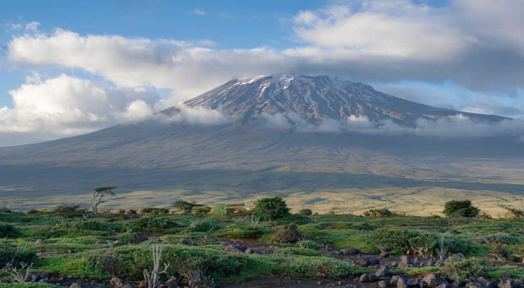 A photo of Mount Kilimanjaro in Tanzania, taken from the base. The mountain is covered with snow at its peak, and its terrain is rocky and barren. There are clouds in the sky, and the ground is covered with green vegetation. The background reveals a vast landscape with rolling hills.