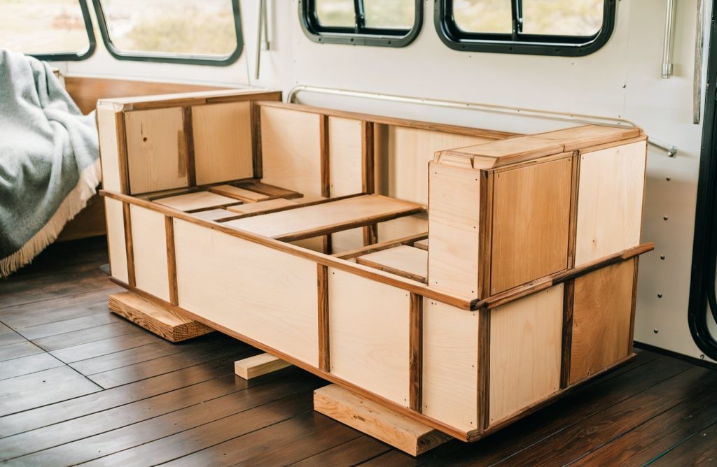 RV furniture, wooden frame sofa under construction. A draped fabric is seen on the left side of the frame. The interior of the RV has a modern aesthetic with a combination of wooden and metallic elements.