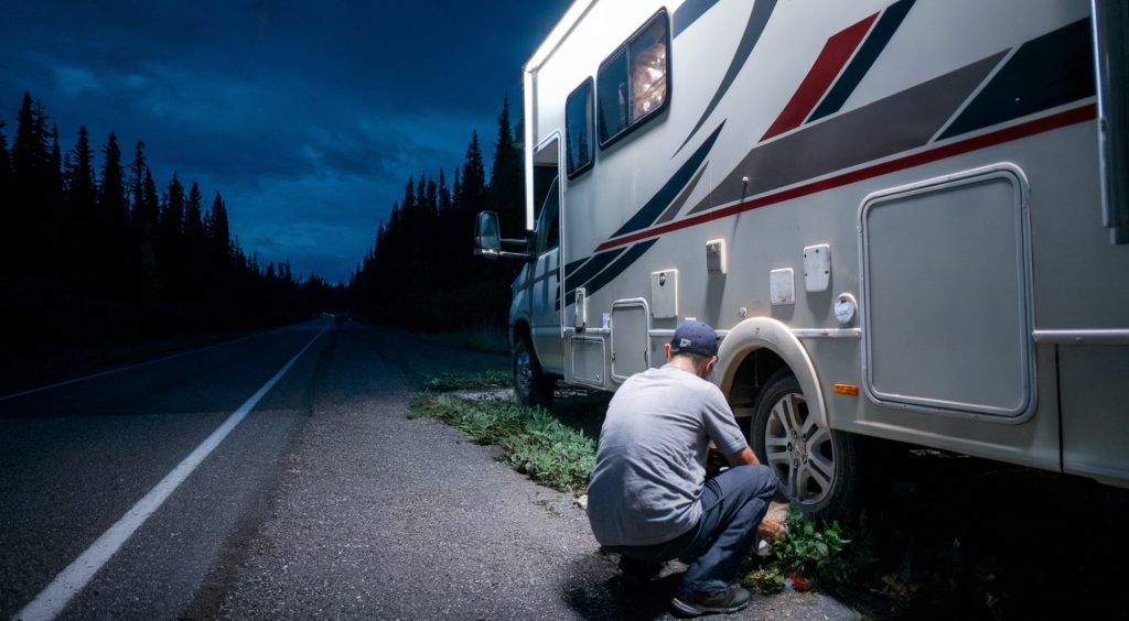 A photo of an RV parked on the side of a dark, one-lane road. The owner, wearing a baseball hat, is seen from behind without tools as he tries to figure out what to do.  The background is a dense forest with tall trees.