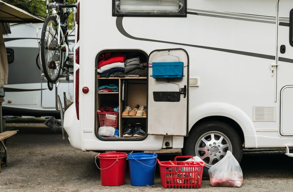 The rear end of a white RV with its door open. Various items are stored inside the RV, including folded clothes, shoes, and other miscellaneous items. Outside the RV, on the ground, there are several objects: two red buckets, a blue bucket, a red basket, and a clear plastic bag. A bicycle is mounted on the RV's exterior, and there's a glimpse of another vehicle in the background.