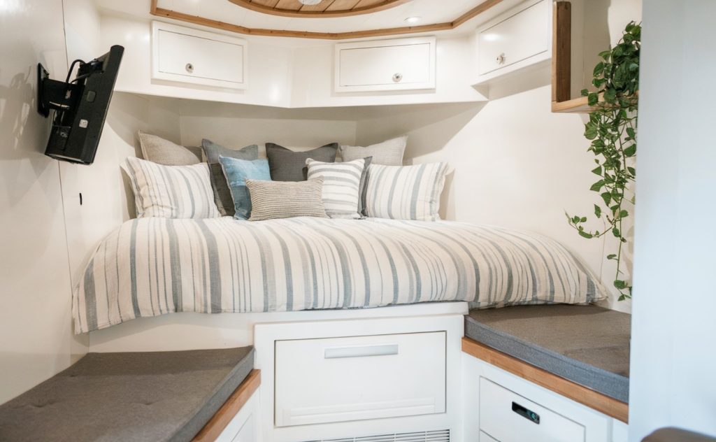 A renovated RV bedroom. To the left, a mounted television is visible, and to the right, there's a wooden shelf with a plant. The room is painted in a light color, and there are white drawers and cabinets below the bed to maximize storage. The overall ambiance is calm and inviting.