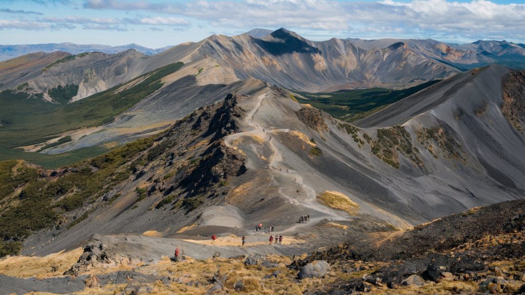 A photo of the Tongariro Alpine Crossing in New Zealand. The image is taken from a distance, showing the trail winding through the mountains. There are hikers on the trail, and the mountains are covered with volcanic rock formations. The sky is clear, with a few clouds.