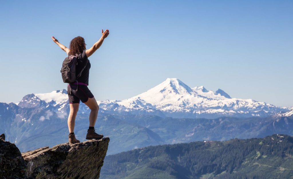 Woman standing on the edge of a cliff raising her hands in triumph for conquering one of the best hiking trails in the world.