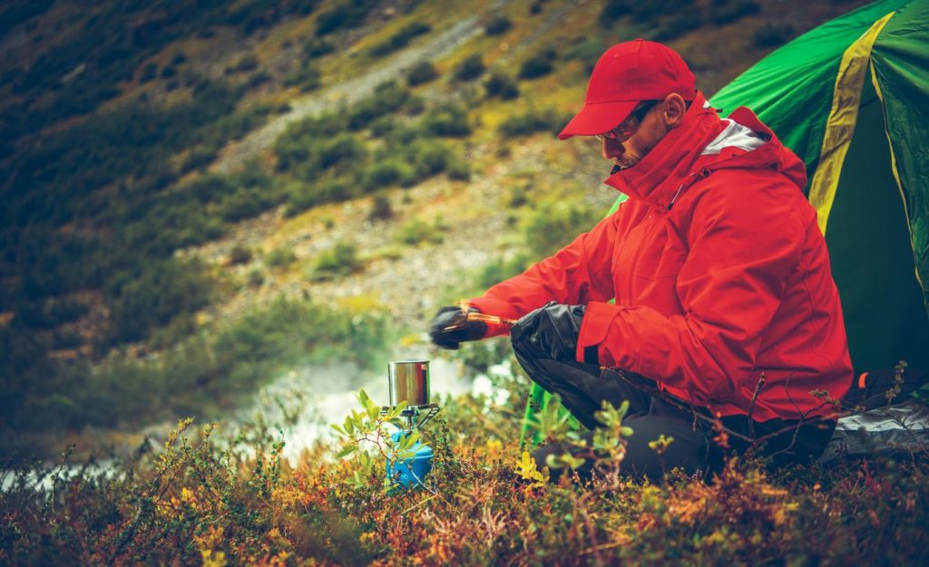 A man in a red outdoor jacket sitting by his tent preparing edible plants over a cooking stove, showing the connection between foraging and outdoor survival.