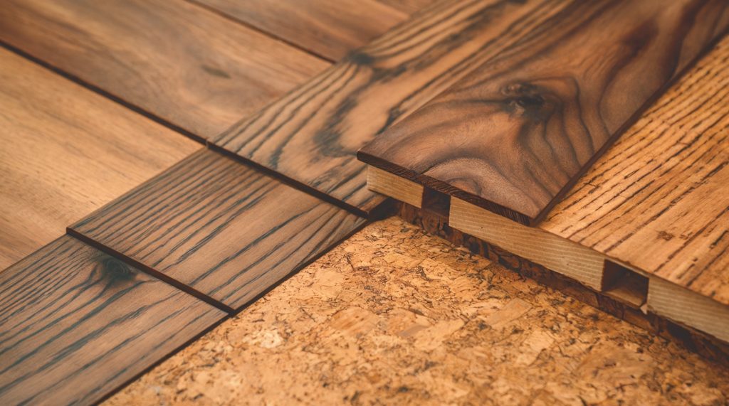 A close-up view of wooden floor planks and a cork surface. The wooden planks have a rich brown hue with grain patterns, and they are laid out in a staggered manner. Adjacent to the wooden planks is a cork surface, which has a textured, beige appearance. The cork seems to be used as a base or underlayment for the wooden planks.