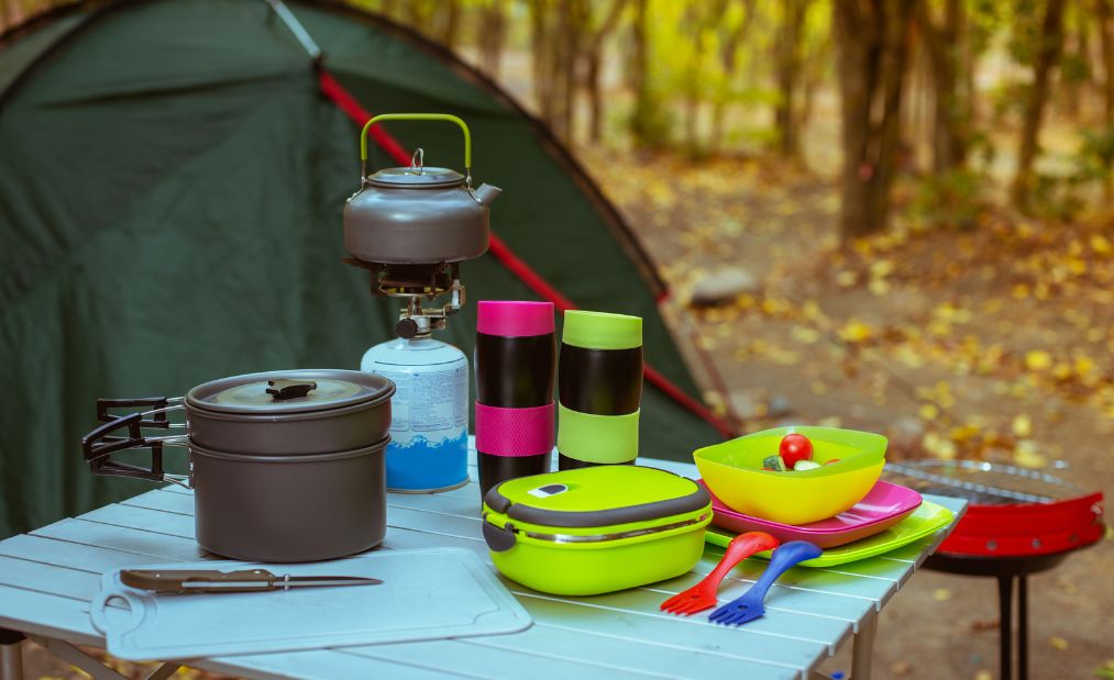 Lightweight camping cookware next to a tent in a forest setting, including collapsible silicone bowls, a backpacker stove and utensils.