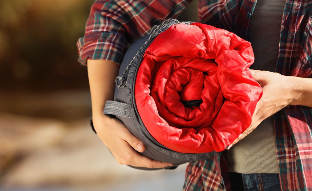 Camper in a plaid shirt carrying a rolled up red sleeping bag.