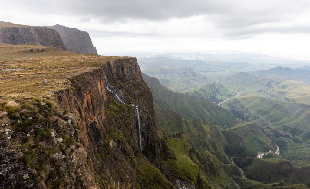 A lone photographer is seen standing on a ridge above Tugela Falls in Africa with a stunning view of the surrounding mountains.