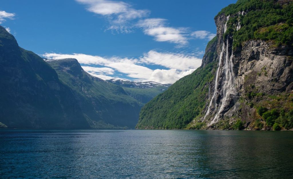 A view of the Seven Sisters Waterfall in Norway from the fjord with green mountains in the distance. 
