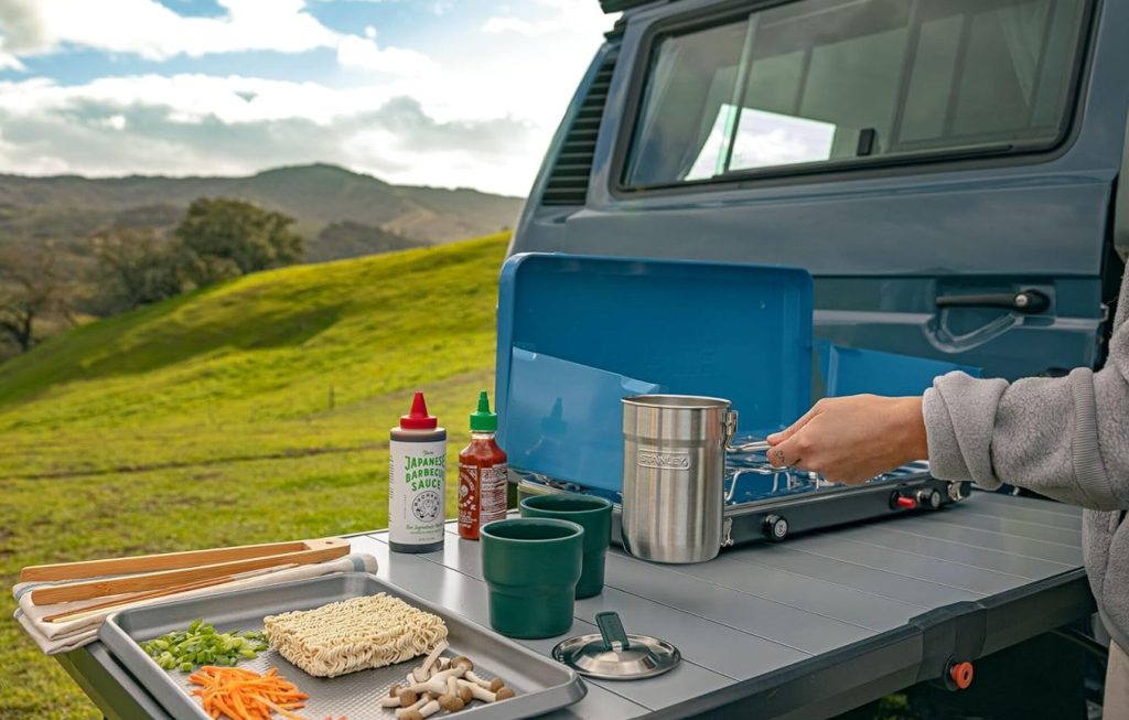 The Stanley Wildfare compact camping cookset seen on a camping table with a portable stove outside the rear of a van. 