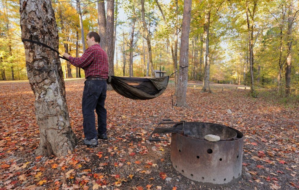 A man in a red flannel plaid shirt is seen employing one of the key knots needed to safely and securely hang a hammock.