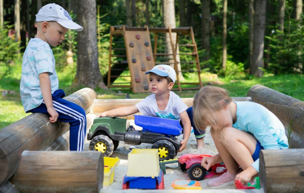 Three kids playing with toys while outdoors in a camping environment. There is a play structure behind them with rock climbing and other activities.
